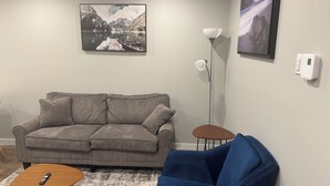 A living room with a grey sofa, a blue armchair, a small wooden coffee table, and a remote control on it. - 2-Bedrooms | On site Parking | near YQR Airport (Regina)