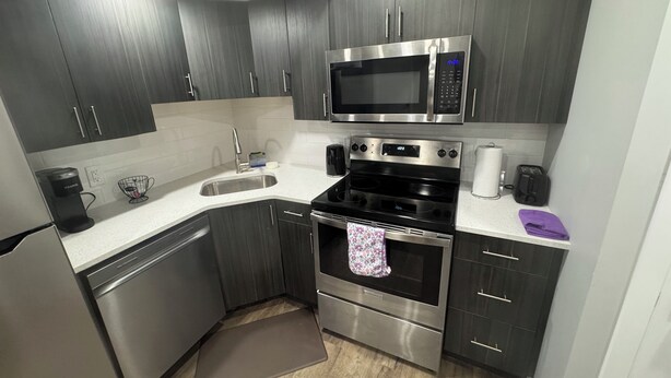 A modern kitchen with dark wooden cabinets, stainless steel appliances including a microwave, oven, and dishwasher, and a white countertop. - 2-Bedrooms | On site Parking | near YQR Airport (Regina)