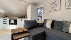 A modern kitchen with white cabinets, a built-in oven, and a coffee maker. There is a grey sofa with cushions and a small wooden table with two plates. - Stay Angels - Cosy Boho Apartment (Sheffield)