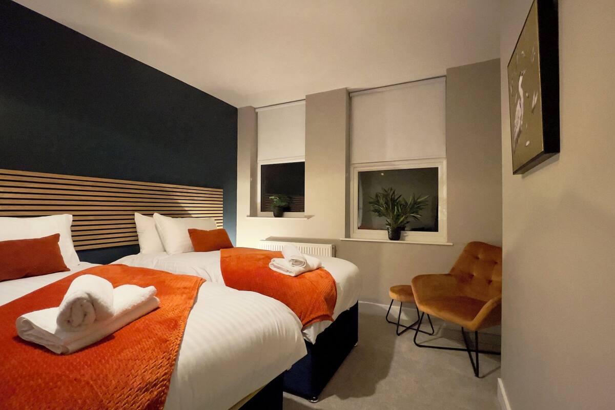 A modern hotel room with a large bed, two bedside tables, a chair, and a window with plants.