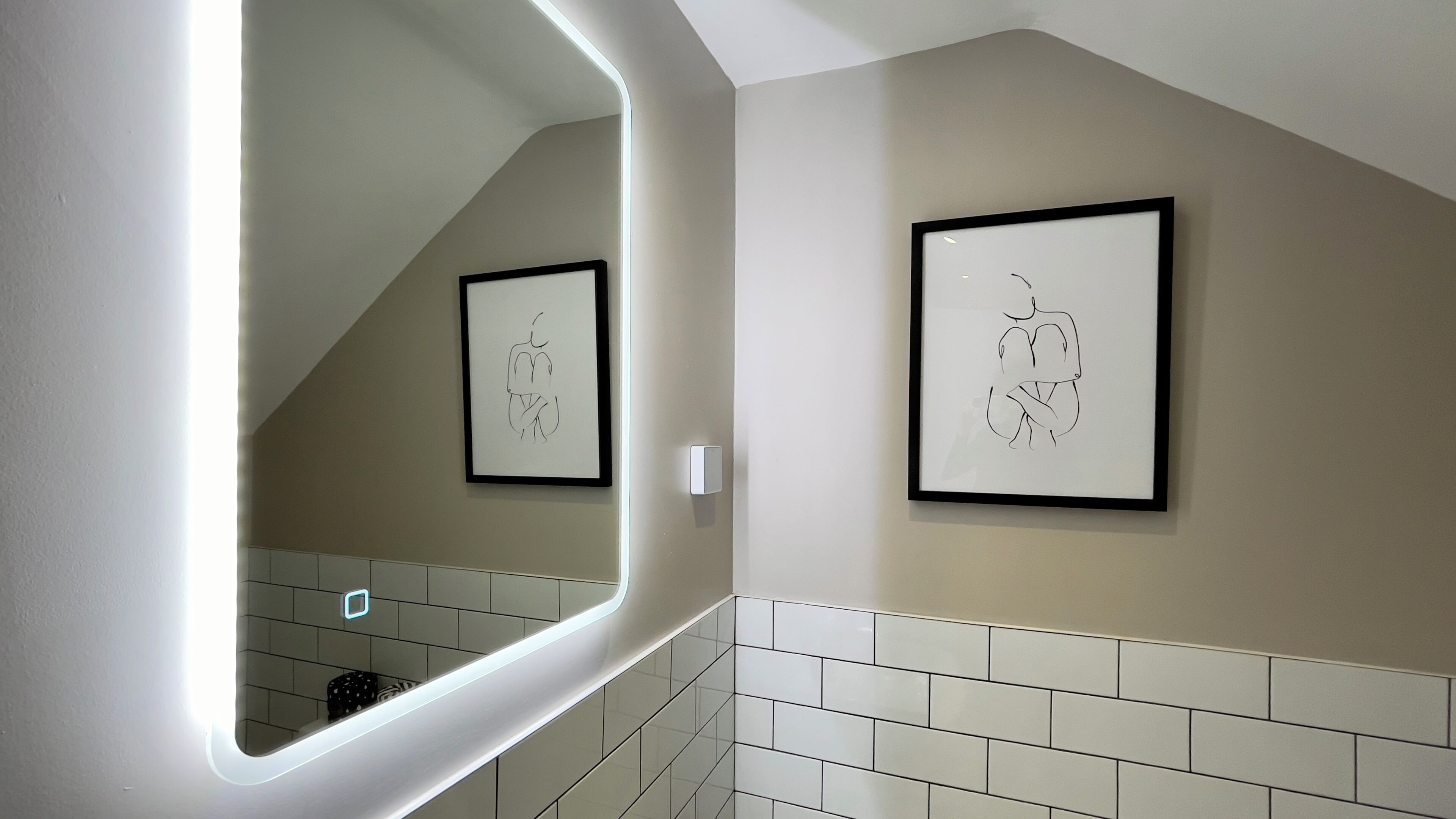 A bathroom with white tiles, a mirror, and two framed line drawings on the wall.