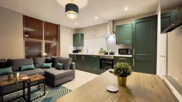 A modern kitchen with green cabinets, a dining table, and a sofa with cushions. - Stay Angels - Cosy Boho Apartment (Sheffield)