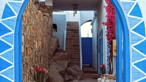 Un chemin de pierre mène à un escalier situé sous une arche bleue, avec un mur en pierre et une chaise en osier.