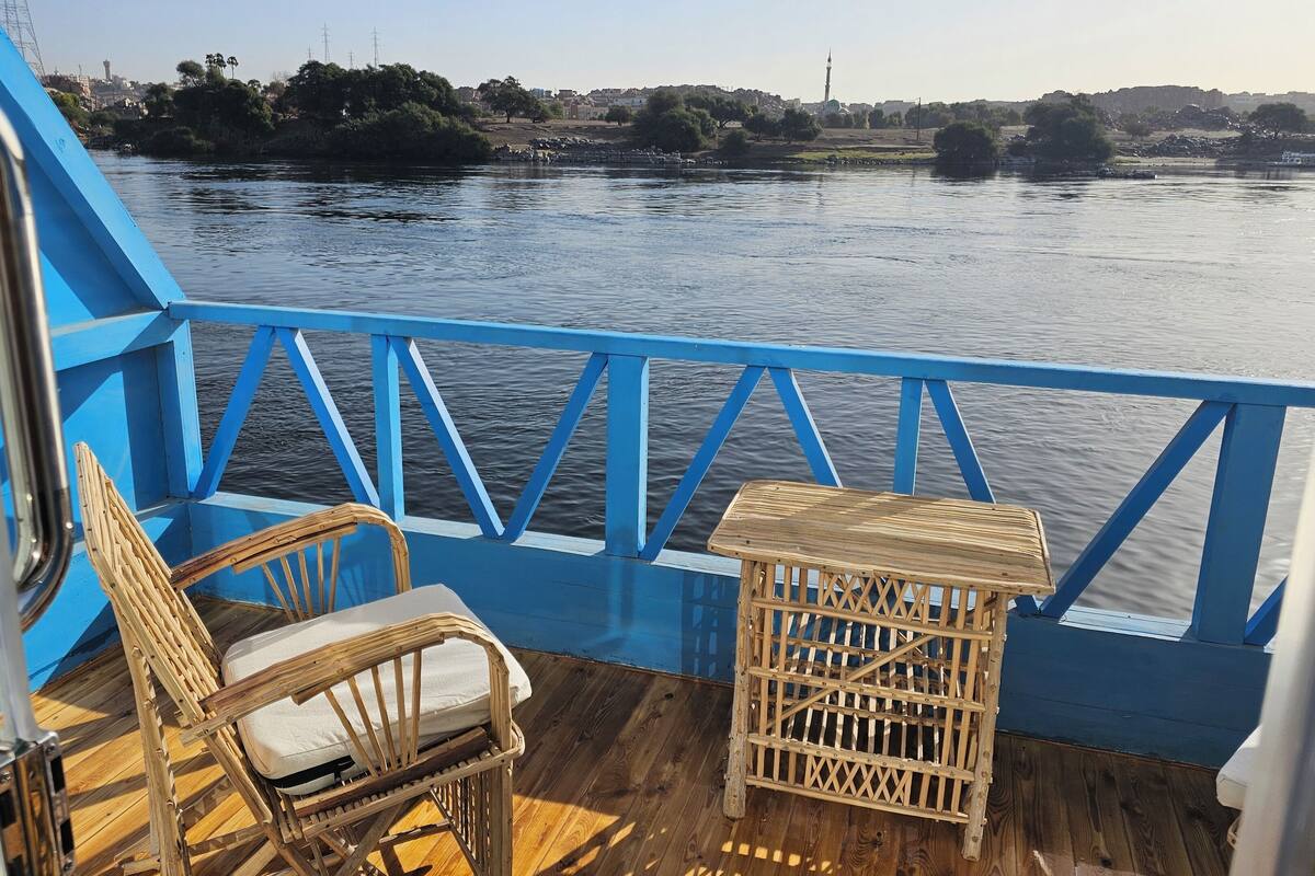 A deck with a blue railing, a wicker chair, a small wooden table, and a view of a river with buildings and trees in the background.