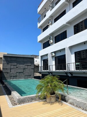 Un edificio moderno con piscina en la azotea, rodeado de una terraza de madera y un muro de piedra.