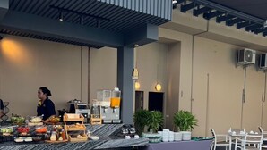 Ein Essbereich im Freien mit gedeckten Tischen, ein Buffet mit verschiedenen Speisen und ein Mitarbeiter, der das Buffet betreut.
