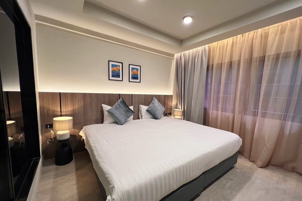 A modern hotel bedroom with a large bed, two bedside lamps, and a view of the outdoors through sheer curtains.