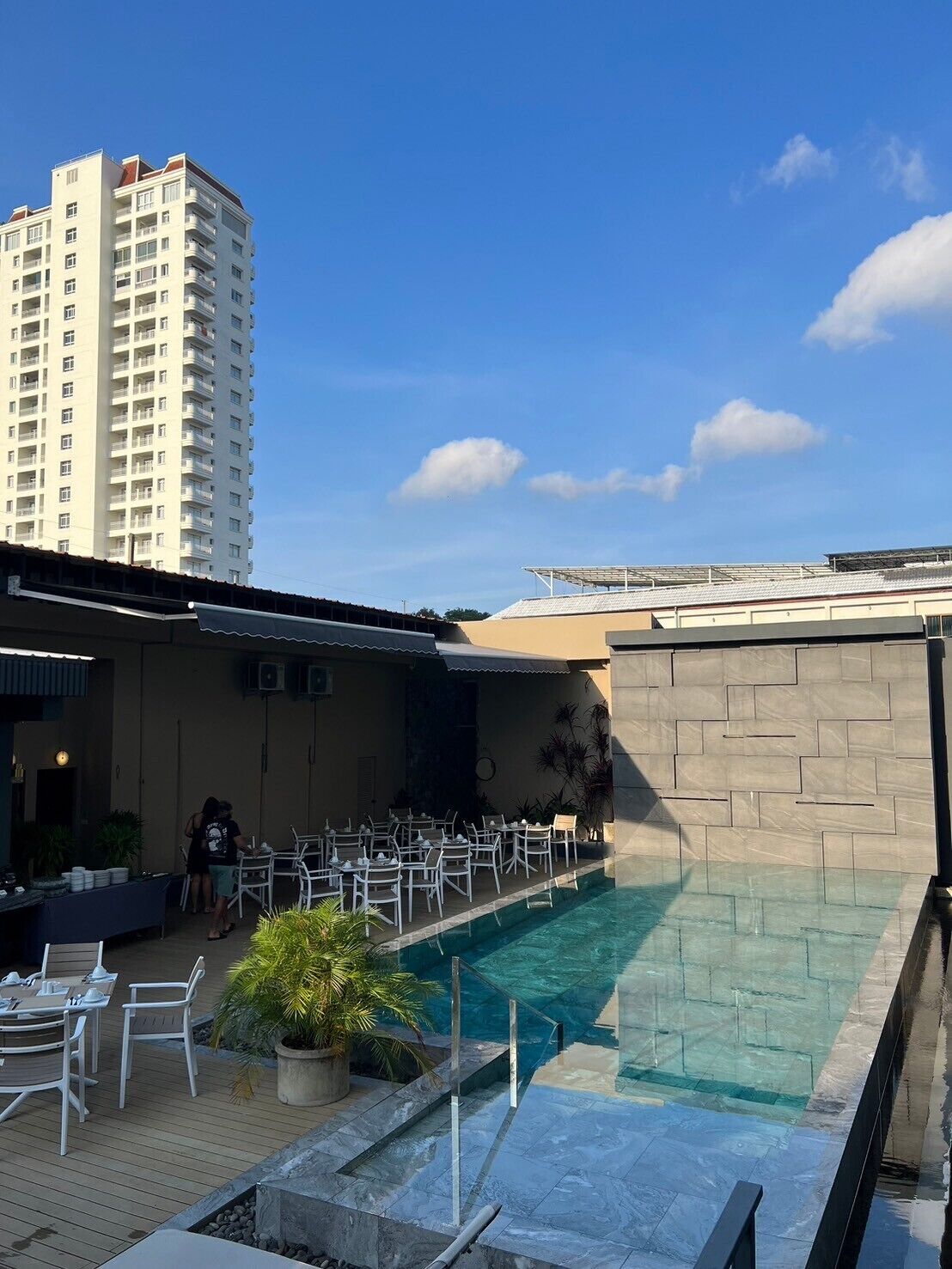 A rooftop pool area with lounge chairs and a dining setup, surrounded by a high-rise building.