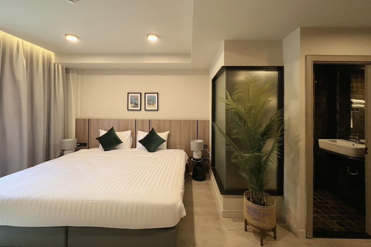 A hotel room with a large bed, two bedside tables, a potted plant, a bathroom with a sink, and two framed pictures on the wall.