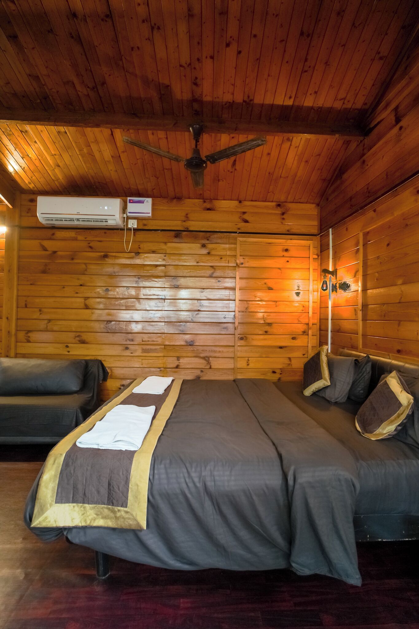 A wooden cabin room with a bed, two sofas, a ceiling fan, and an air conditioning unit.