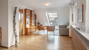 Ein modernes Wohnzimmer mit Holzboden, einer integrierten Bücherwand, einer Essecke mit Stühlen, einem Sofa und einem großen Fenster mit Aussicht.