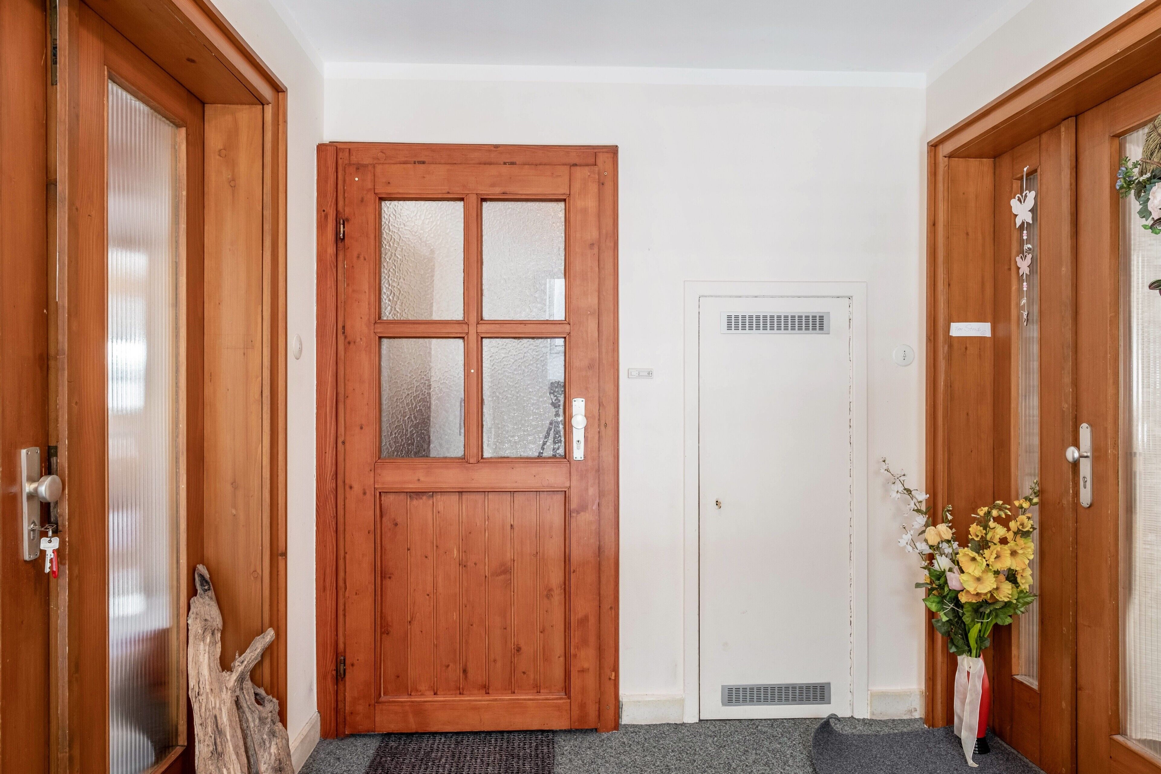 Eine Holztür mit Glasfenster, eine weiße Tür mit Lüftungsgitter und eine Vase mit Blumen.
