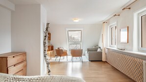 Ein Schlafzimmer mit einem Holzkoffer, einem Bett mit Quilzdecke, einem Fenster mit Aussicht, einem Sessel und einem Heizkörper.