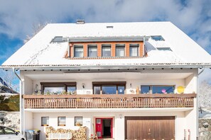 A two-story building with a red door, a balcony with a wooden railing, and a garage. - Apartment '5, 82 Qm' with Shared Garden and Wi-Fi (Lenzkirch)