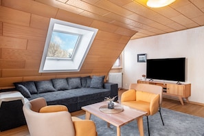 A modern loft-style room with a sloped ceiling, a large window, a flat-screen TV, a sofa, and a coffee table.