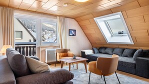 A cozy living room with a sofa, armchair, and a table. There is a window with a view of a snowy landscape. - Apartment '4, 65 Qm' with Shared Garden, Balcony and Wi-Fi (Lenzkirch)