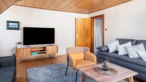A living room with a flat-screen TV, wooden entertainment unit, sofa, armchair, and a coffee table with a vase. - Apartment '4, 65 Qm' with Shared Garden, Balcony and Wi-Fi (Lenzkirch)