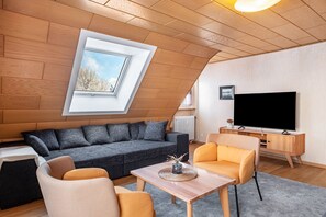 Une pièce de style loft avec un plafond en pente, une grande fenêtre, un canapé gris, des fauteuils orange, une table basse en bois et un téléviseur posé sur un meuble en bois.