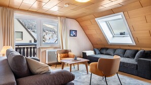 Ein gemütliches Wohnzimmer mit einer Couch, einem Sessel und einem kleinen Tisch, ein Fenster mit Blick auf verschneite Bäume und ein Oberlicht.