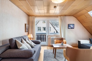 Une pièce confortable avec un canapé, une table et une chaise. Il y a une fenêtre avec des rideaux et une vue sur les bâtiments à l’extérieur.
