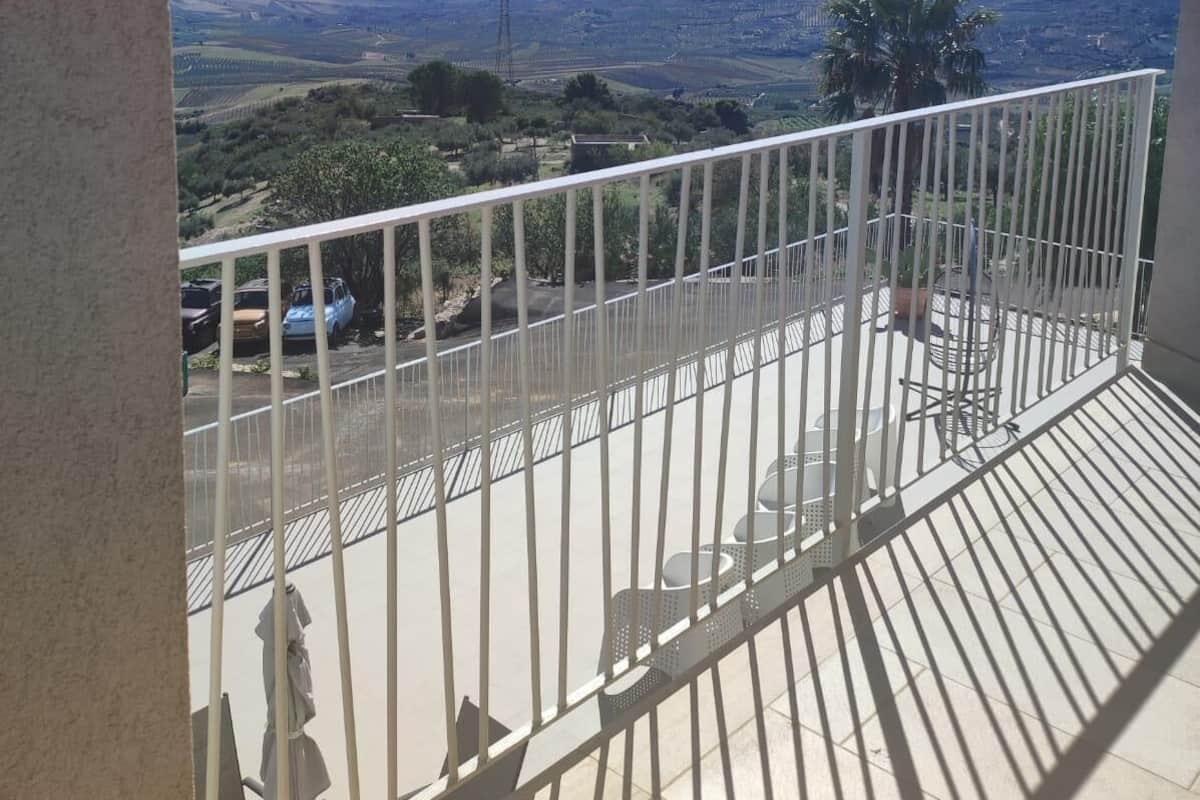 A balcony with a metal railing, a view of a hilly landscape, and parked cars in the background.
