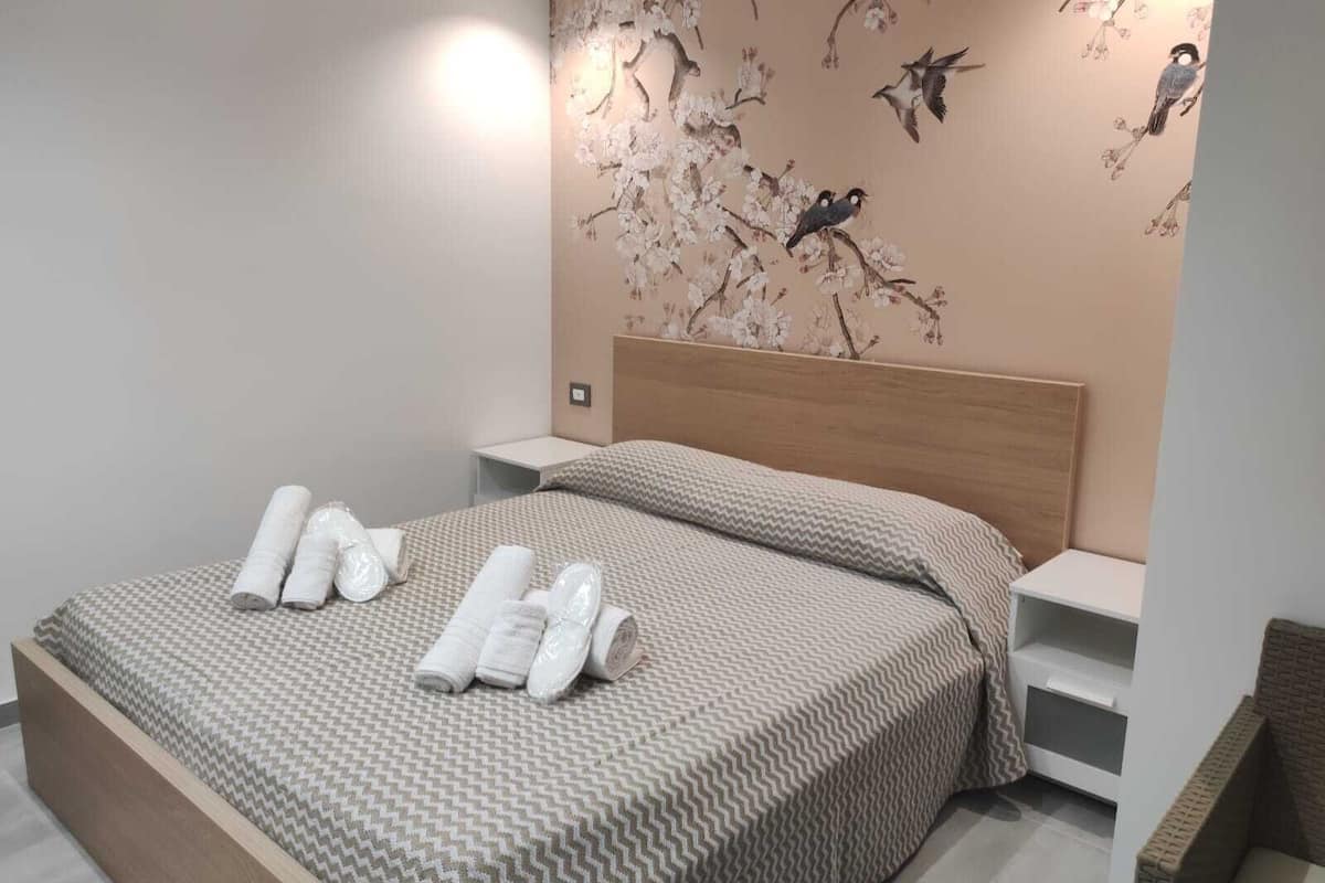 A neatly made bed with a patterned bedspread and pillows, a wall with a floral mural, and a small bedside table with a lamp.