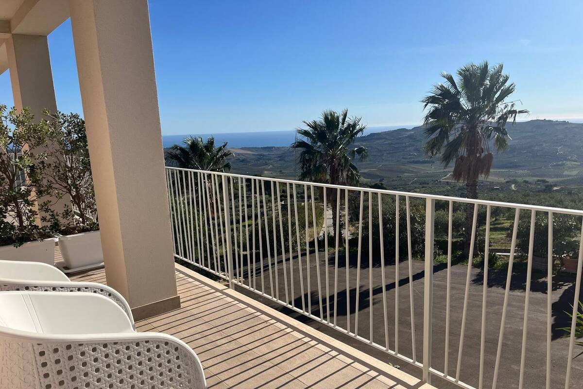 A balcony with a white railing overlooking a scenic landscape with palm trees and distant hills.