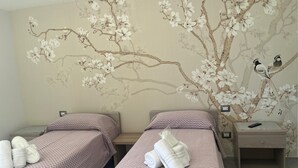 A hotel room with two beds, each with a pink and white patterned bedspread, and a wall mural of a tree with flowers and birds. - TENUTE LAZZARINO (Sciacca)