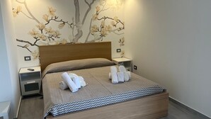 A modern bedroom with a wooden headboard, a bed with a patterned bedspread, and a wall mural of a tree with flowers. - TENUTE LAZZARINO (Sciacca)