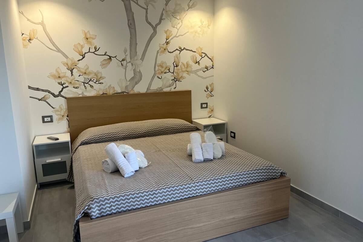 A modern bedroom with a wooden headboard, a bed with a patterned bedspread, and a wall mural of a tree with flowers.
