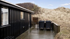A wooden deck leading to a black cabin with two chairs and a table, surrounded by tall grass and a clear sky.