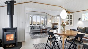 A modern interior with a wood-burning stove, a dining area with a round table and chairs, a sofa, and a large window offering a view of the outdoors.
