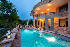 Un espace luxueux aménagé autour d’une piscine, avec un bâtiment aux lignes courbes, des sièges d’extérieur et une ambiance chaleureuse grâce à un éclairage soigné.
