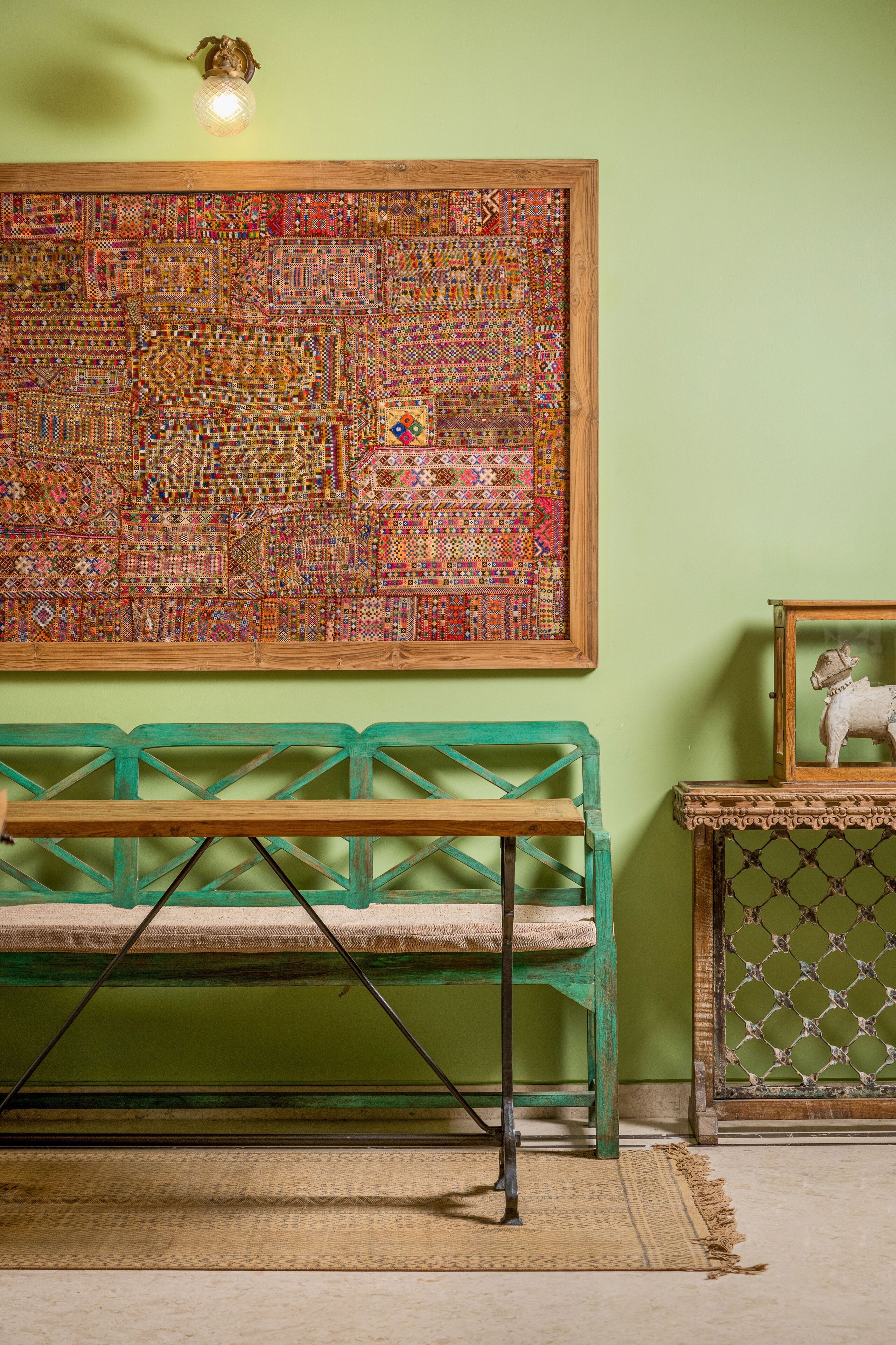 A room with a large colorful tapestry on the wall, a green bench, and a small table with a decorative item.