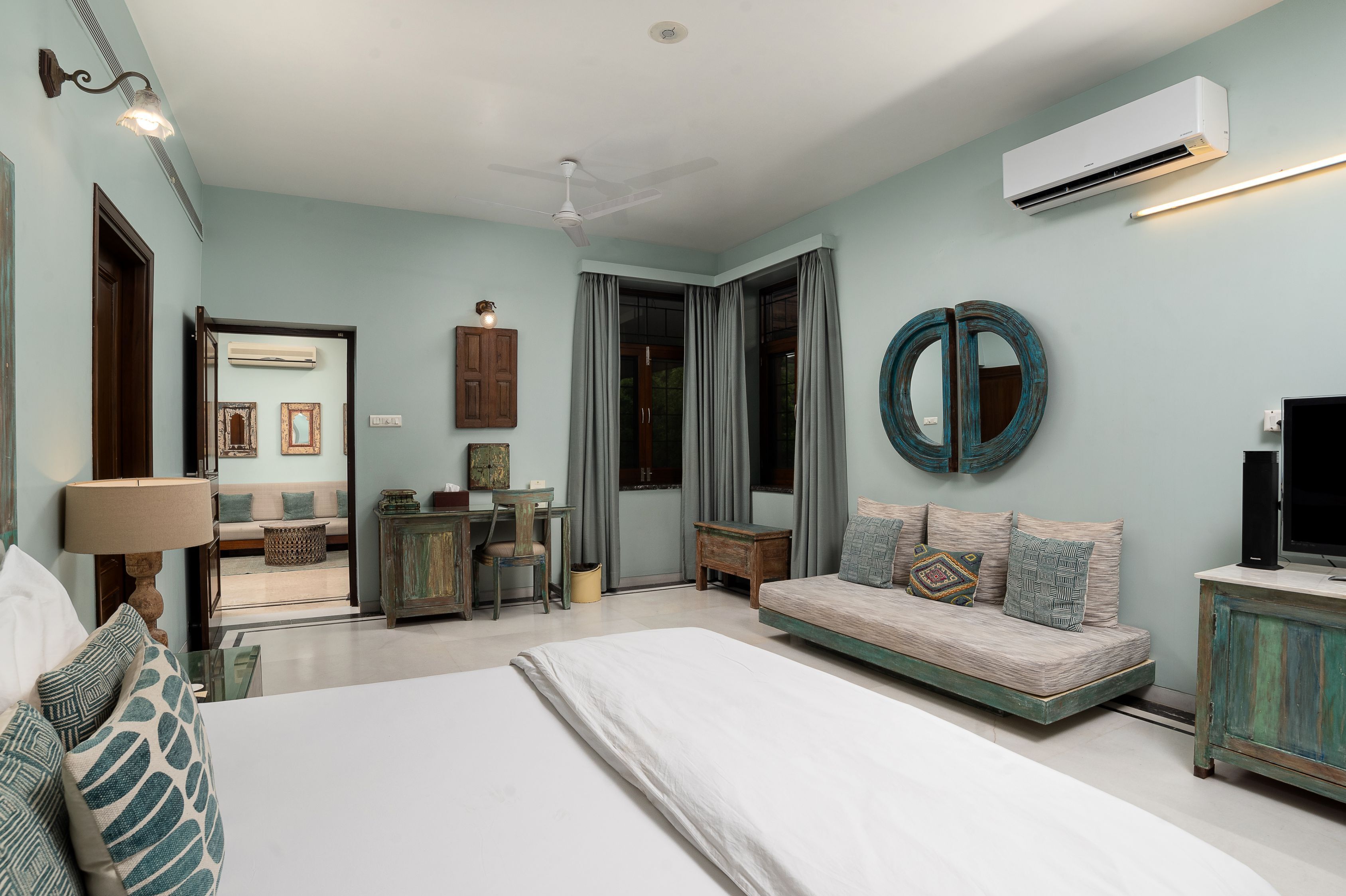 A bedroom with a bed, a desk, a sofa, a mirror, and an air conditioning unit.