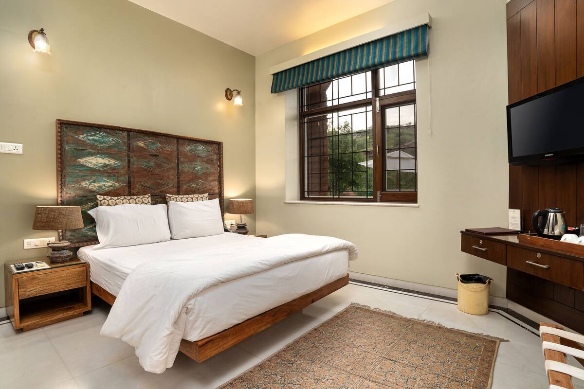 A bedroom with a large bed, wooden headboard, bedside lamps, a television, and a window with a view of greenery.