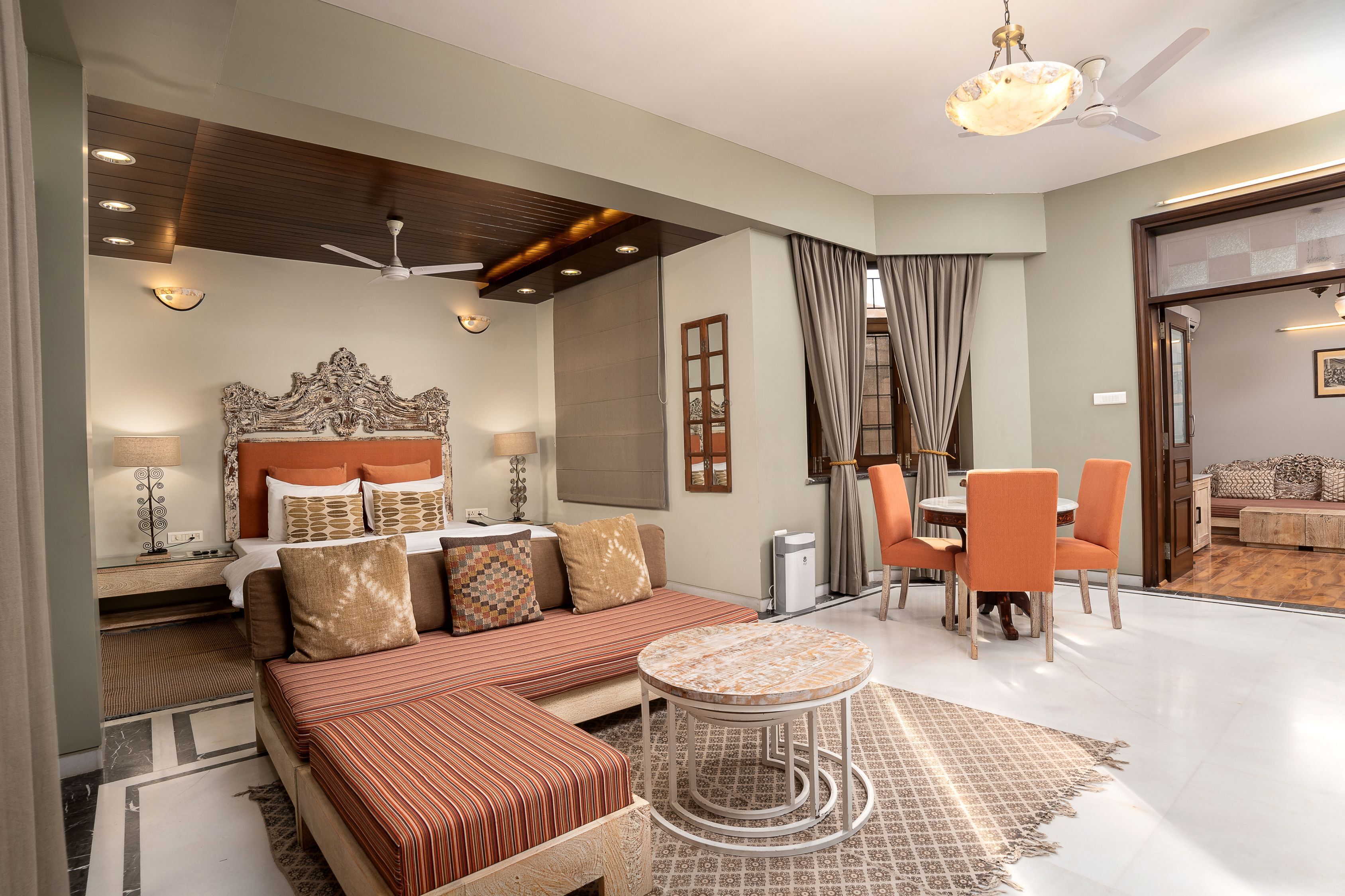 A modern hotel room with a large bed, a sofa, a dining area with orange chairs, and a ceiling fan.