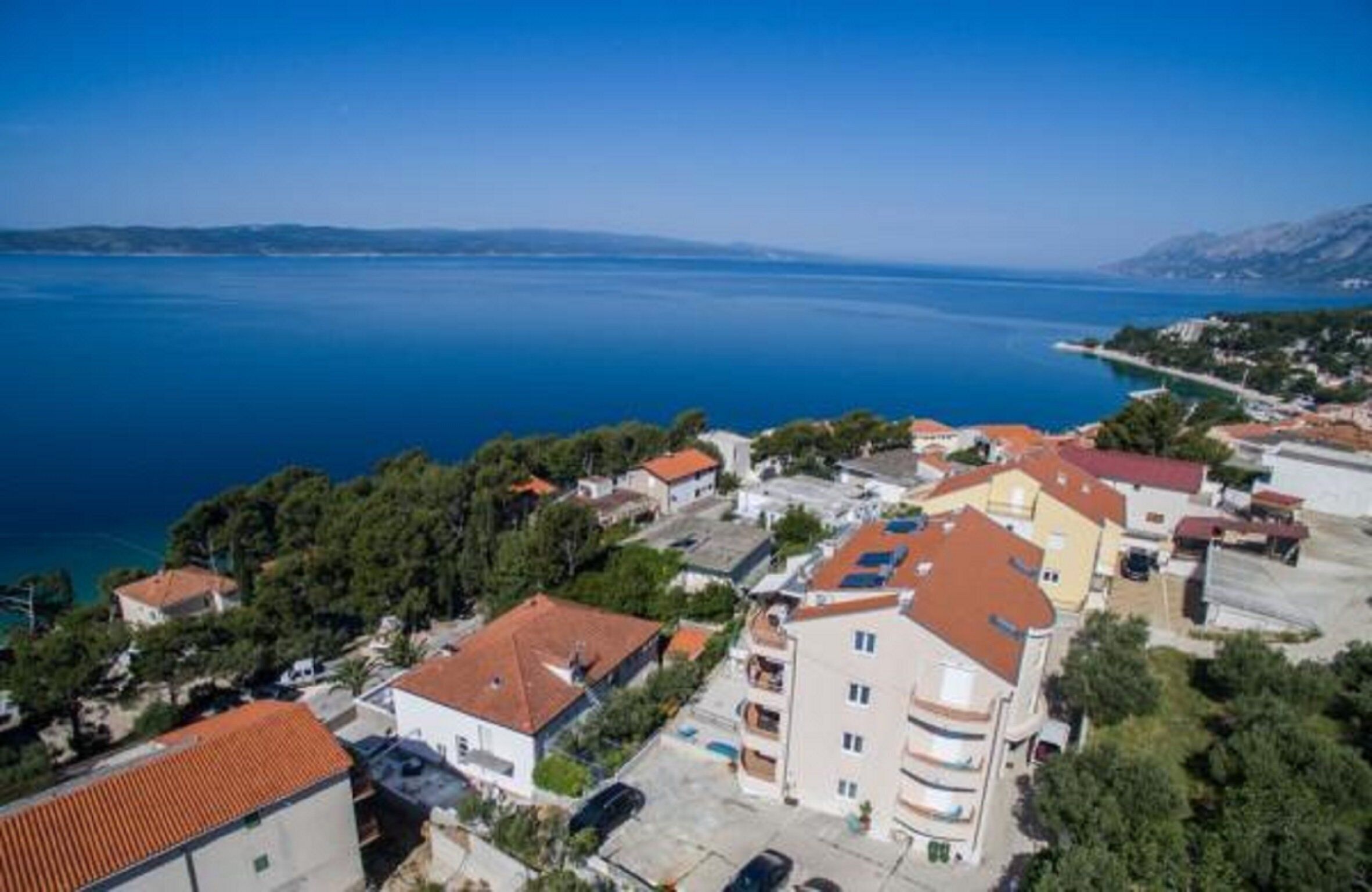 A coastal residential area with houses, a clear blue sea, and a hilly landscape in the distance.