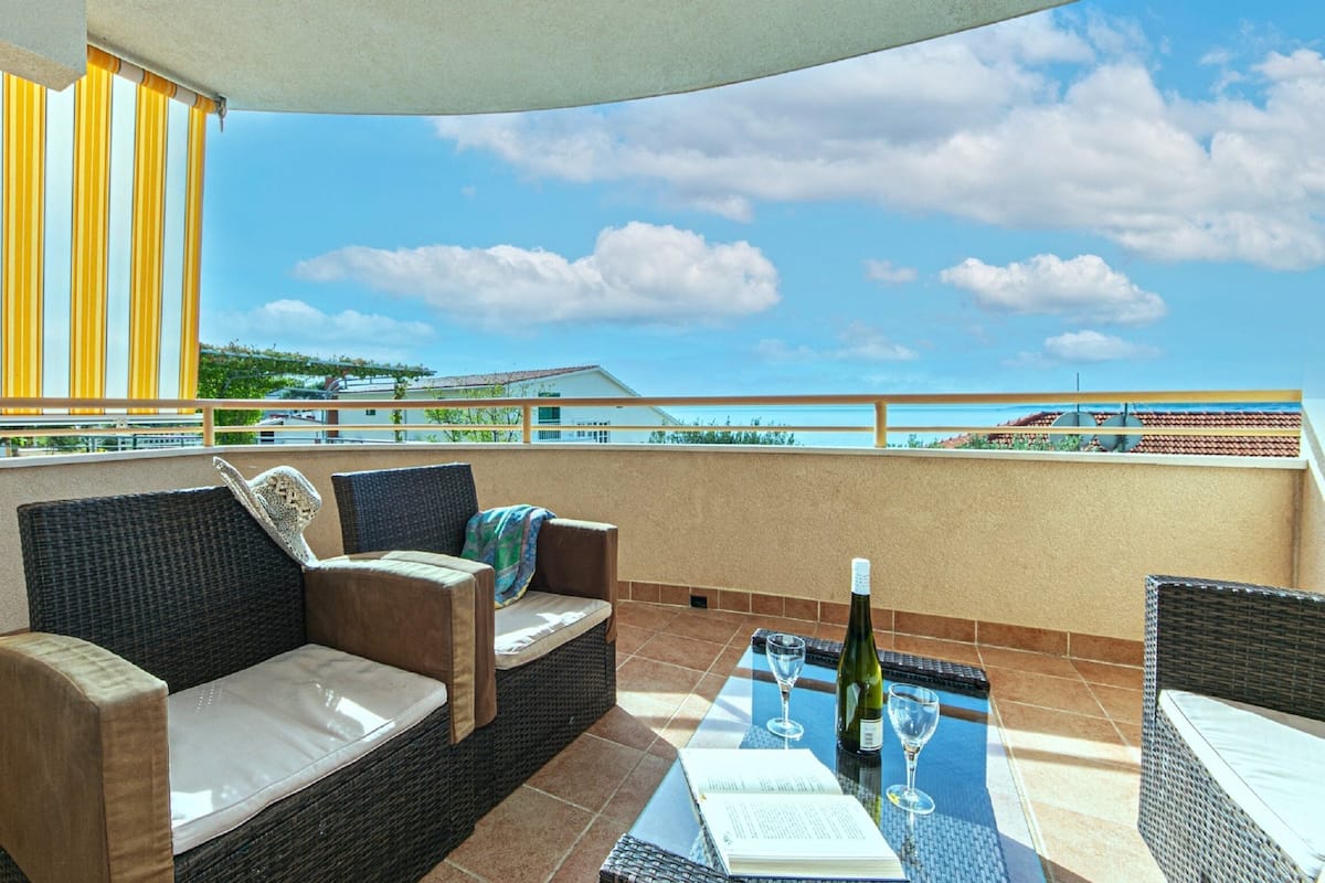 A balcony with wicker furniture, a table with a bottle and glass, and a view of buildings in the distance.