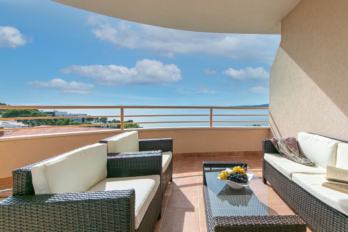 A balcony with wicker furniture, a glass table with fruit, and a view of the sea.