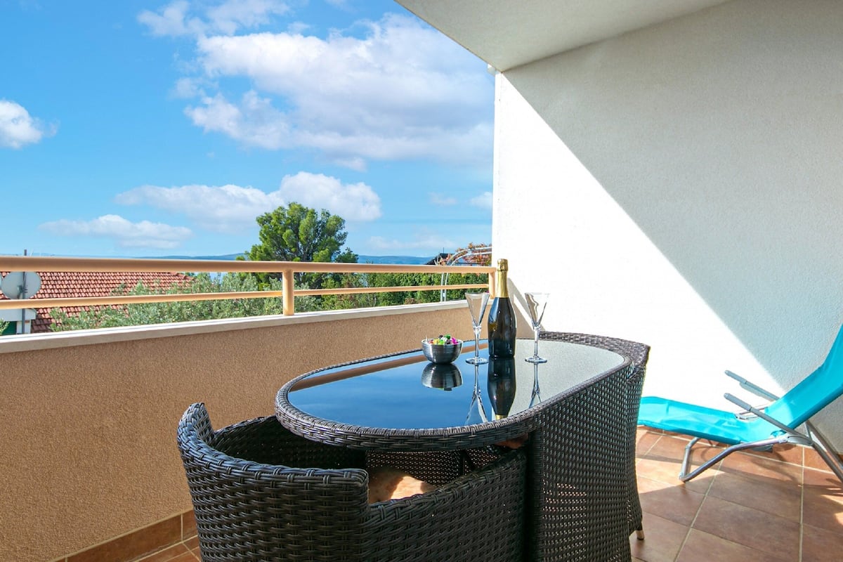 A balcony with a wicker table, a blue chair, and a view of the sea and mountains.