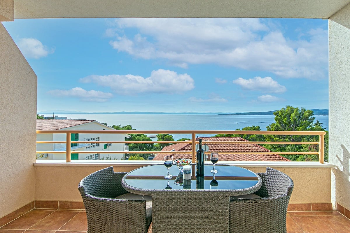 A balcony with a table and chairs, overlooking a coastal view with buildings and trees.