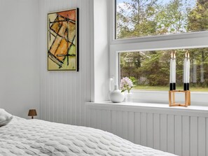 Un dormitorio con cama, ventana con vista a árboles, un cuadro en la pared y un florero con flores.