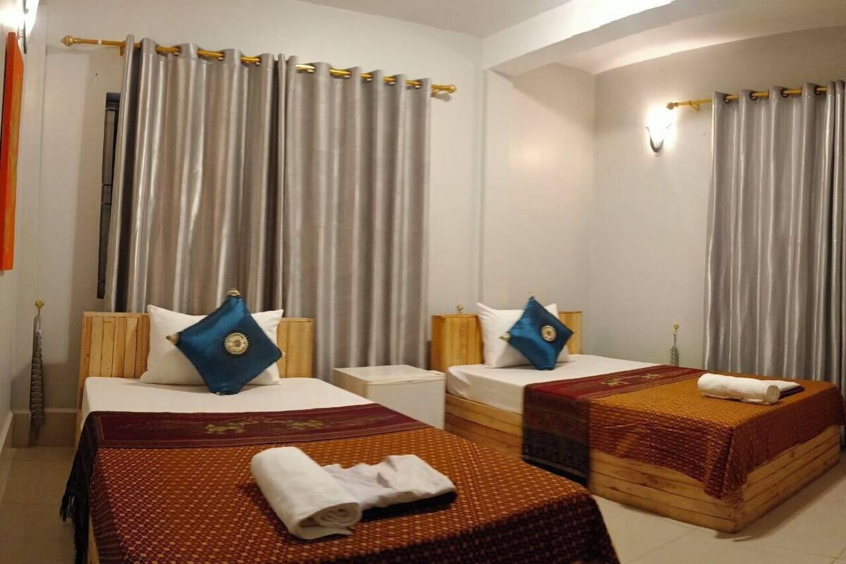 Ein Hotelzimmer mit zwei Betten, jedes mit einer rot-weißen Bettdecke, einem blauen Kopfkissen und einem Holz-Kopfteil. Zwischen den Betten steht ein kleiner, weißer Nachttisch, und am Fußende der Betten eine hölzerne Bank.