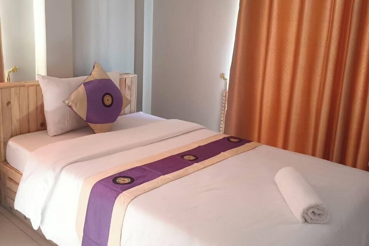 Ein ordentlich gemachtes Bett mit weiß-lila Bettdecke, einem Holzkopfstück und einem braunen Vorhang.