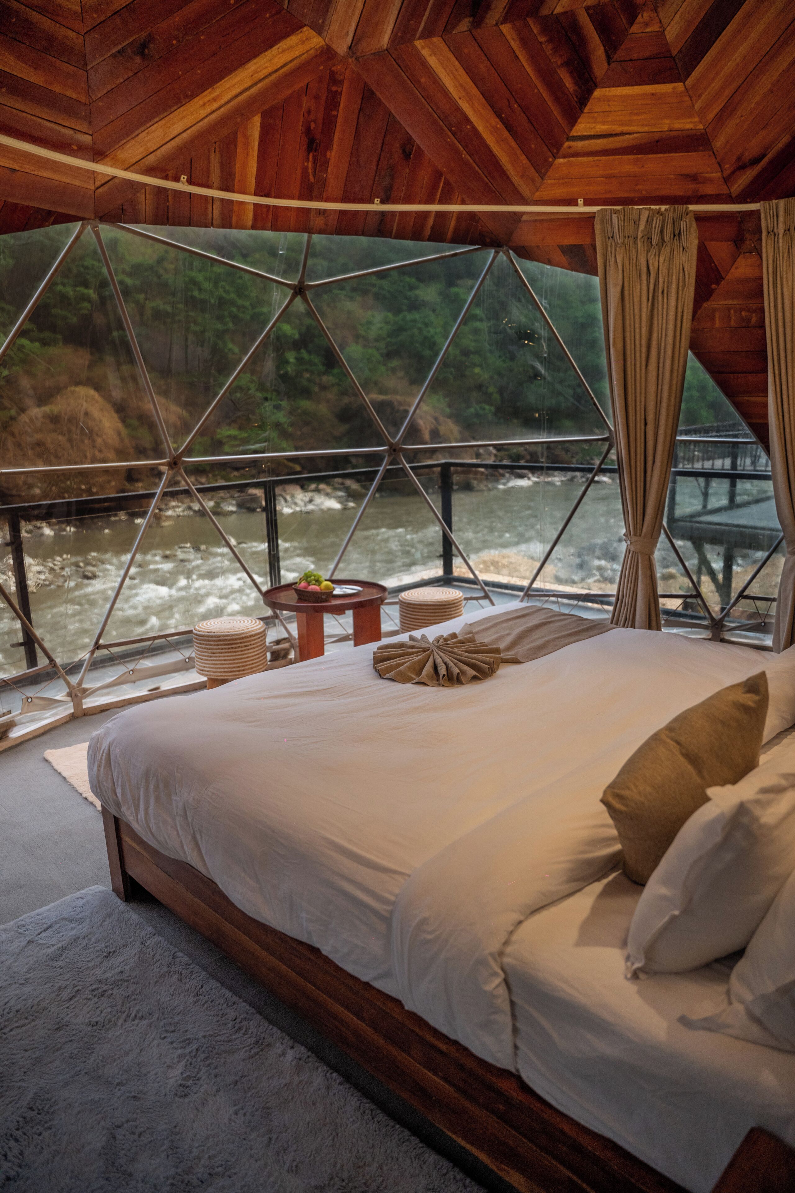 A bed with a view of a river, wooden ceiling, and a window with curtains.
