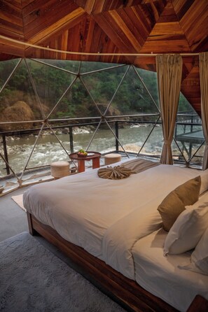Una cama con vista a un río, techo de madera y una ventana con cortinas.