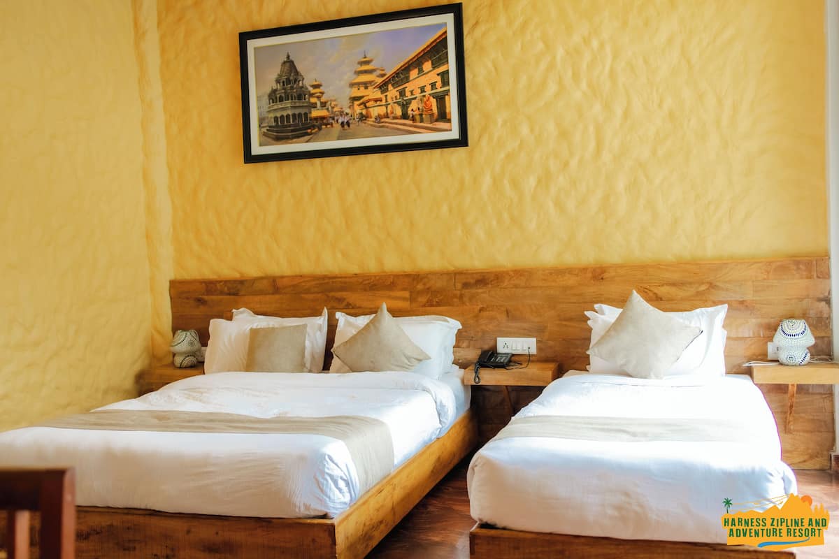 A hotel room with two beds, wooden headboards, a framed picture on the wall, a bedside table with a lamp, and a small clock.
