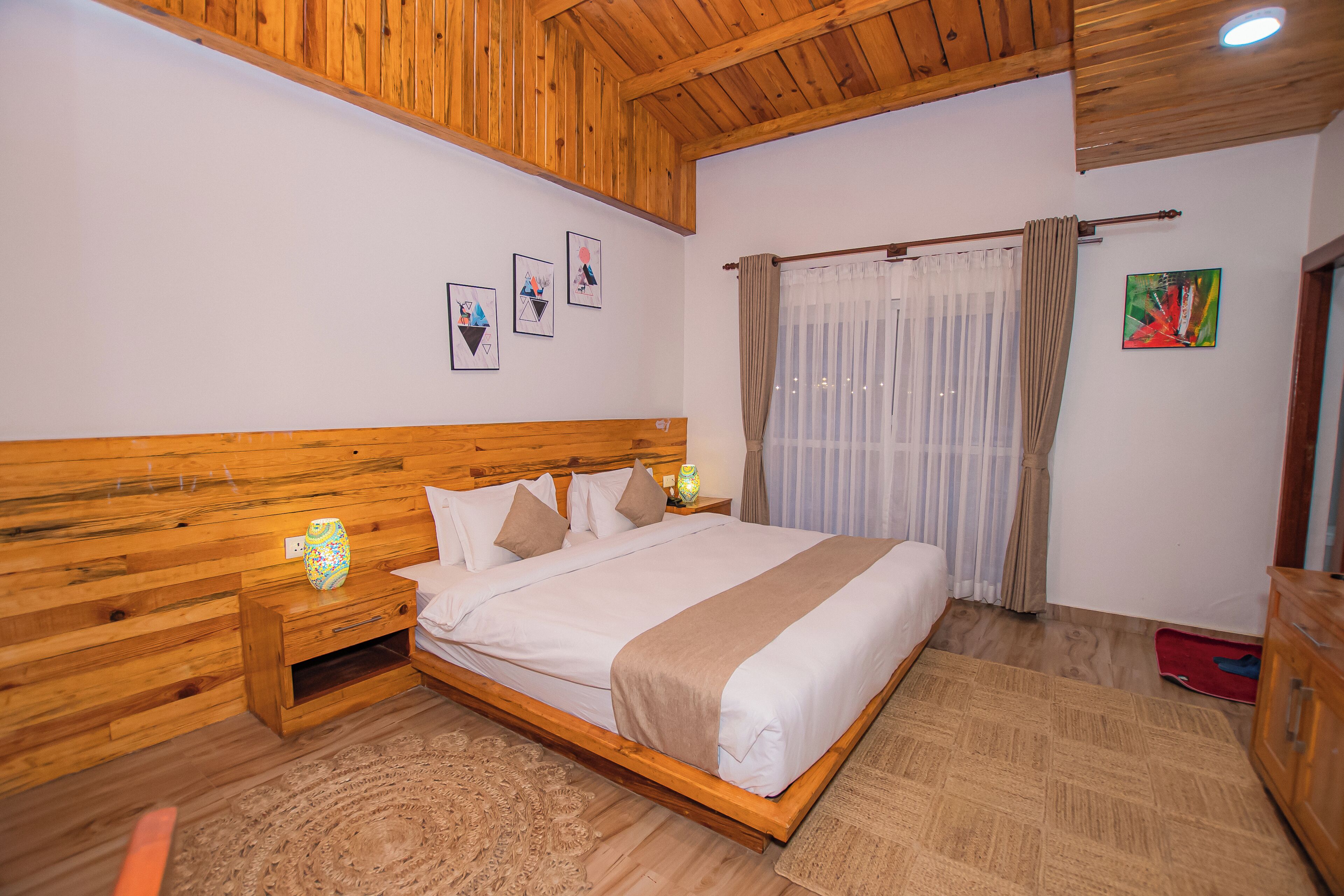 Una cama con estructura de madera, un edredón y almohadas color beige, una mesita de noche con una lámpara y un piso de madera.