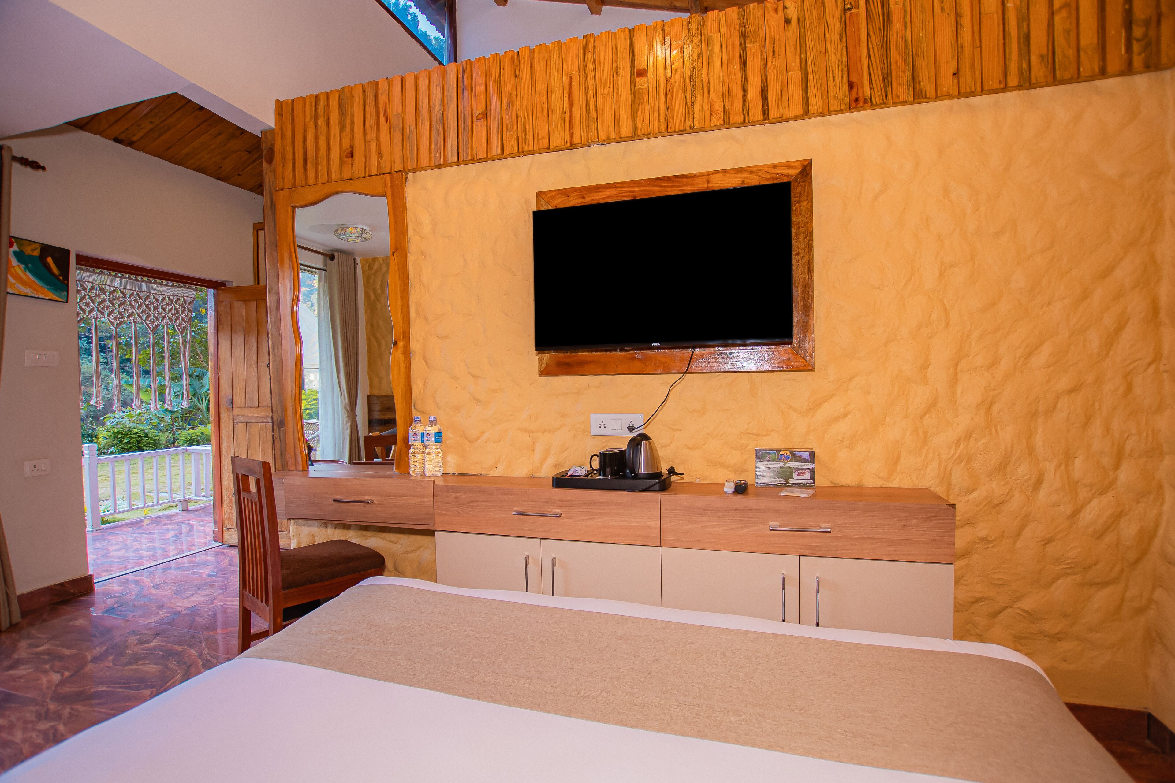 Quarto com cabeceira de madeira, cama, cómoda de madeira, televisão na parede e janela com vista para vegetação.